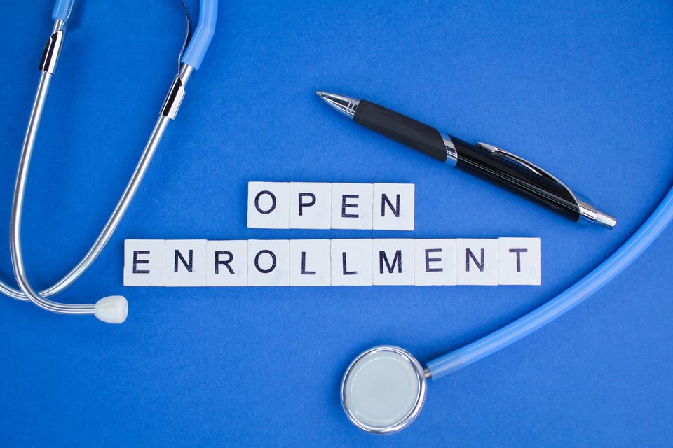 A stethoscope, a pen and lettered tiles spelling OPEN ENROLLMENT arranged on a blue background.