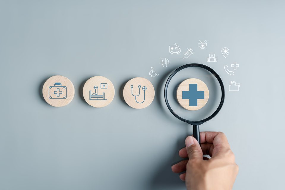 A hand holds a magnifying glass over a row of wooden discs printed with health-care icons, with more icons drawn above them.