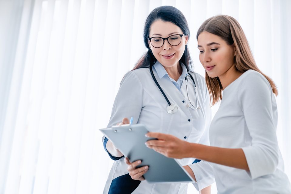 Two women stand together reading a clipboard, one in a white coat with a stethoscope, in a bright room with vertical blinds.