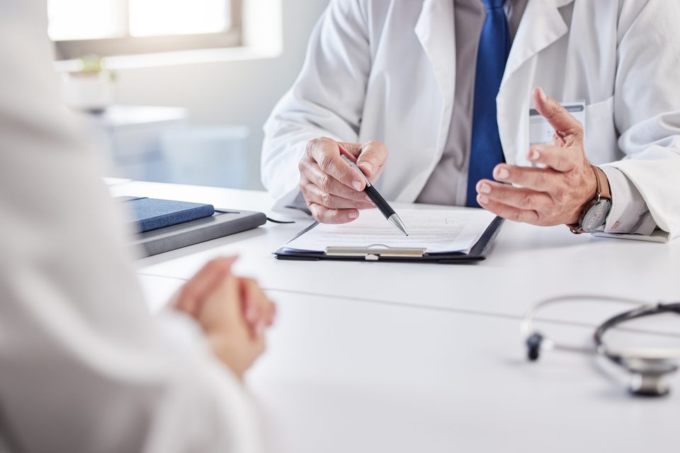 Across a white desk, a person in a white coat points a pen at a clipboard while talking with someone seated opposite; a stethoscope rests on the desk.