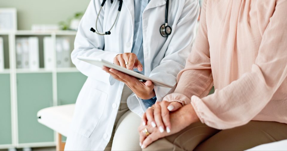 A clinician in a white coat holds a tablet beside a seated person in a pink blouse, in a consulting room with shelved binders.