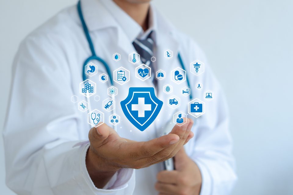 A clinician holds an open palm beneath a large illustrated shield with a cross, surrounded by a ring of small blue health-care icons.