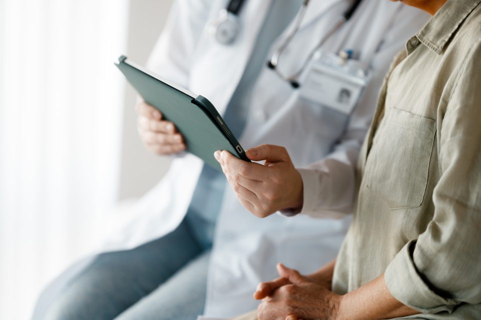 A clinician in a white coat and a seated person in a linen shirt look together at a tablet held between them.