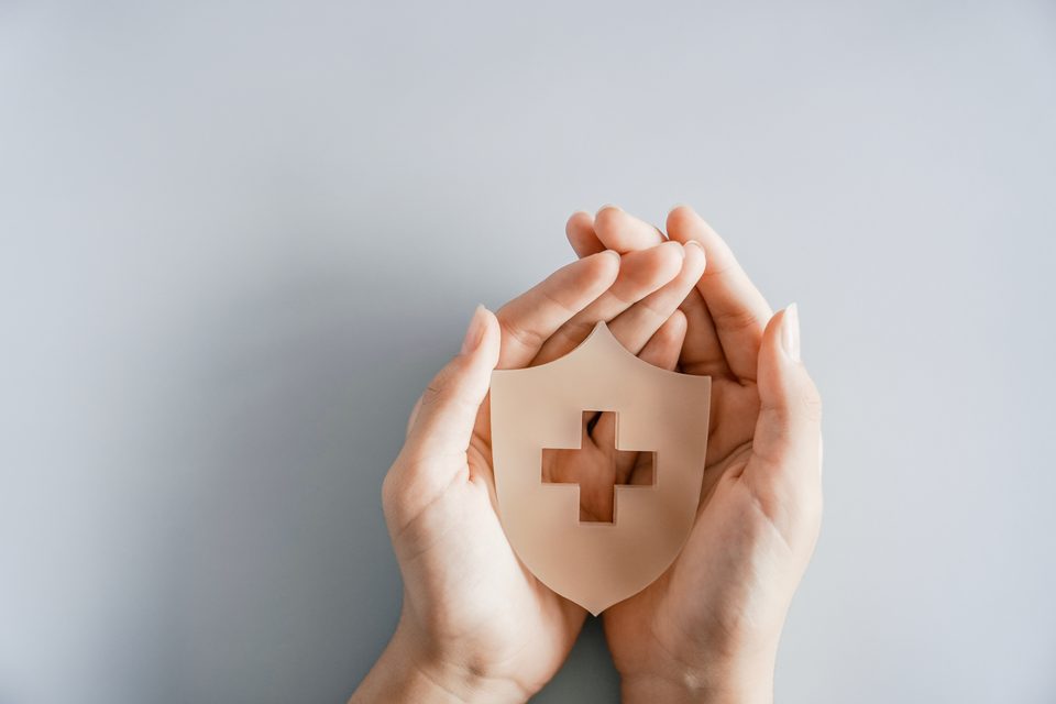Two hands cradle a paper shield cut with a cross, photographed from above on a plain grey background.