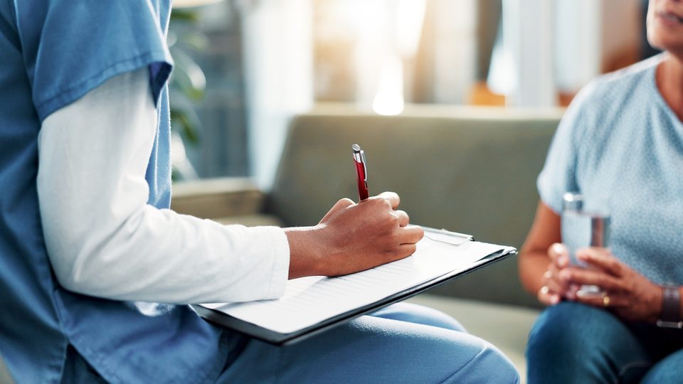 A person in scrubs writes on a clipboard while a seated person holding a glass of water looks on, in a room with a sofa and daylight.