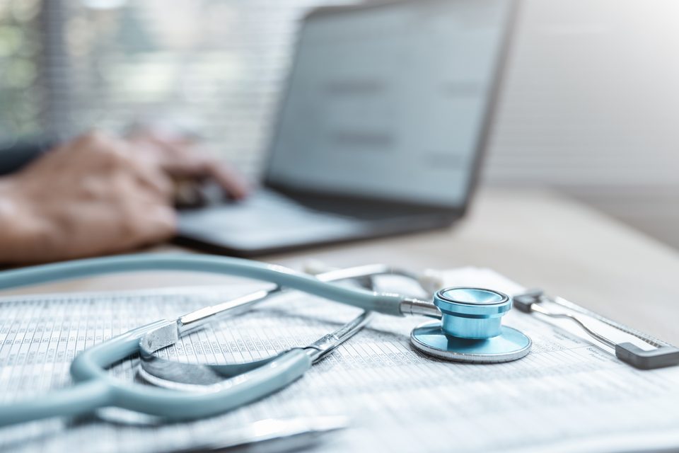 A stethoscope lies across printed record sheets on a desk, with a person typing at a laptop out of focus behind.
