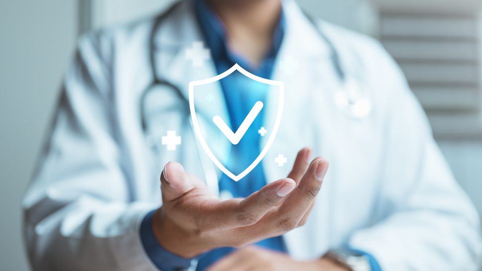 A clinician in a white coat holds an open palm beneath an illustrated shield marked with a check.
